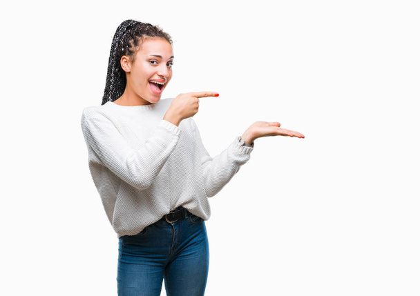 junge geflochtene Haare afrikanische Amerikanerin trägt Winterpullover über isoliertem Hintergrund erstaunt und lächelt in die Kamera, während sie mit der Hand präsentiert und mit dem Finger zeigt. - Foto, Bild