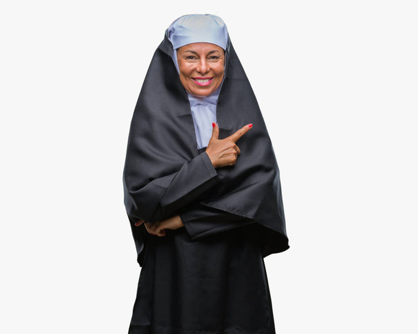 Middle age senior christian catholic nun woman over isolated background cheerful with a smile of face pointing with hand and finger up to the side with happy and natural expression on face looking at the camera. - Photo, image