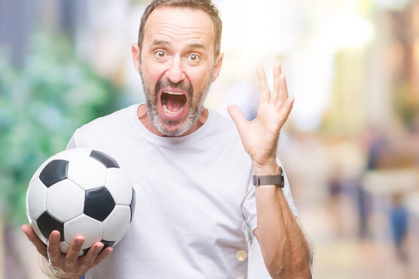 Edad media hoary hombre mayor sosteniendo pelota de fútbol sobre fondo aislado muy feliz y emocionado, expresión ganadora celebrando la victoria gritando con una gran sonrisa y las manos levantadas
 - Foto, Imagen