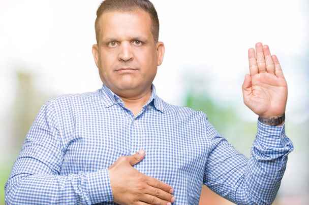 Hombre de negocios árabe de mediana edad sobre un fondo aislado jurando con la mano en el pecho y la palma abierta, haciendo un juramento de lealtad promesa
 - Foto, Imagen