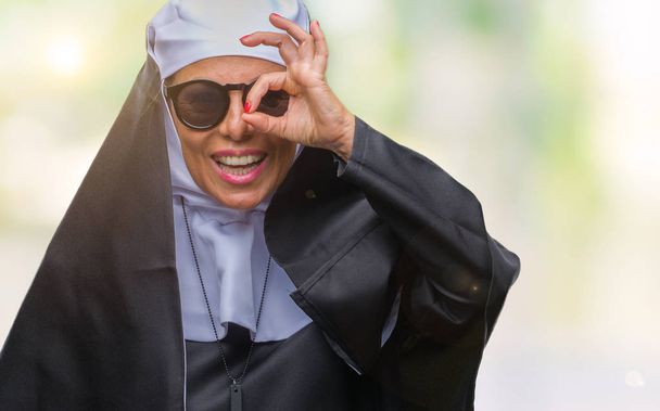 Seniorin der katholischen Nonne mittleren Alters trägt Sonnenbrille über isoliertem Hintergrund und macht ok Geste mit lächelnden Händen, Auge blickt durch Finger mit glücklichem Gesicht. - Foto, Bild