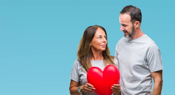Middle age hispanic casual couple in love holding red heart over isolated background with a confident expression on smart face thinking serious - Foto, imagen