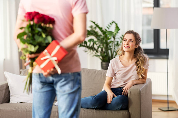 woman looking at man with flowers and gift at home - Фото, зображення
