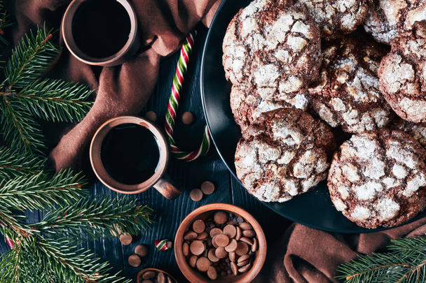 close-up of Christmas Chocolate Crinkle cookies on a plate on a black wooden table with fir tree, candy canes, brown cloth and earthenware cups of coffee, rustic style, view from above, flat lay - Фото, изображение