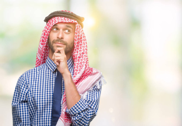 Giovane bell'uomo d'affari arabo che indossa keffiyeh su sfondo isolato con mano sul mento pensando alla domanda, espressione pensierosa. Sorridente con la faccia pensierosa. Concetto di dubbio
. - Foto, immagini