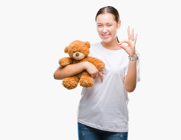 Καυκάσιος γυναίκα κρατώντας αρκουδάκι πάνω από απομονωμένες φόντο κάνει εντάξει πινακίδα με τα δάχτυλα, εξαιρετική σύμβολο - Φωτογραφία, εικόνα