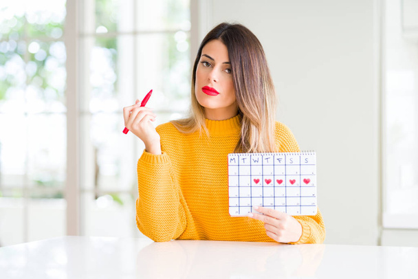 Молода красива жінка тримає календар менструацій вдома з впевненим виразом на розумне обличчя думаючи серйозно
 - Фото, зображення
