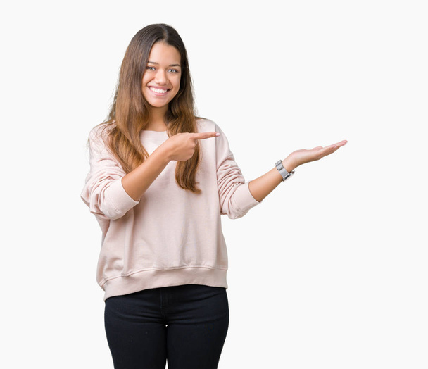 Młoda piękna brunetka kobieta ubrana różowa Bluza na białym tle zdziwiony i uśmiecha się do kamery podczas prezentacji ręką i wskazując palcem. - Zdjęcie, obraz