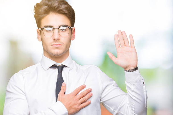 Jovem homem de negócios usando óculos sobre fundo isolado Jurando com a mão no peito e palma aberta, fazendo uma promessa de lealdade juramento
 - Foto, Imagem