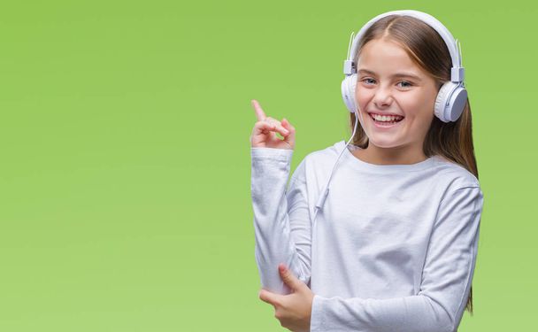 Young beautiful girl wearing headphones listening to music over isolated background with a big smile on face, pointing with hand and finger to the side looking at the camera. - 写真・画像
