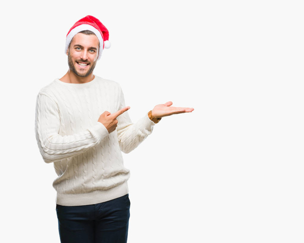 Young handsome man wearing santa claus hat over isolated background amazed and smiling to the camera while presenting with hand and pointing with finger. - Foto, imagen