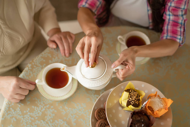 Hands of young woman with white china teapot pouring tea into cup of her senior mother - Фото, зображення