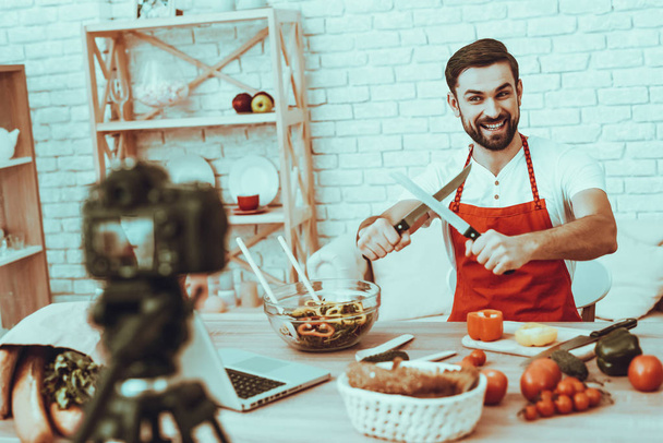 Blogger Makes a Video. Blogger is Smiling Beard Man. Video About a Cooking. Camera Shoots a Video. Laptop and Different Food on Table. Man Showing a Knives. Man in Studio Interior. - Foto, afbeelding