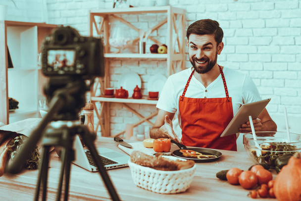 Happy Man Blogger Makes a Video. Video About a Cooking. Camera Shoots a Video. Laptop and Different Food on Table. Man Showing a Pieces of Pepper on Pan. Man Holding a Tablet. Man in Studio Interior. - Foto, Bild