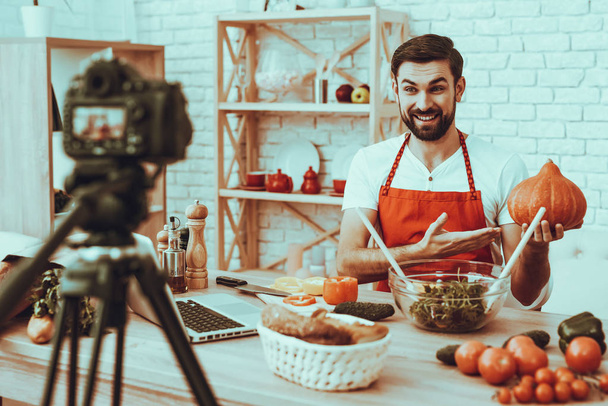 Blogger Makes a Video. Blogger is Smiling Beard Man. Video About a Cooking. Camera Shoots a Video. Laptop and Different Food on Table. Man Showing a Pumpkin. Man in Studio Interior. - Foto, afbeelding