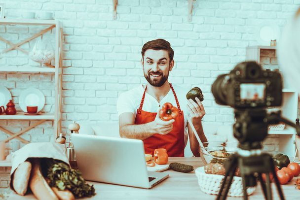 Blogger Makes a Video. Blogger is Smiling Beard Man. Video About a Cooking. Camera Shoots a Video. Laptop and Different Food on Table. Man Showing a Peppers. Man in Studio Interior. - Foto, afbeelding