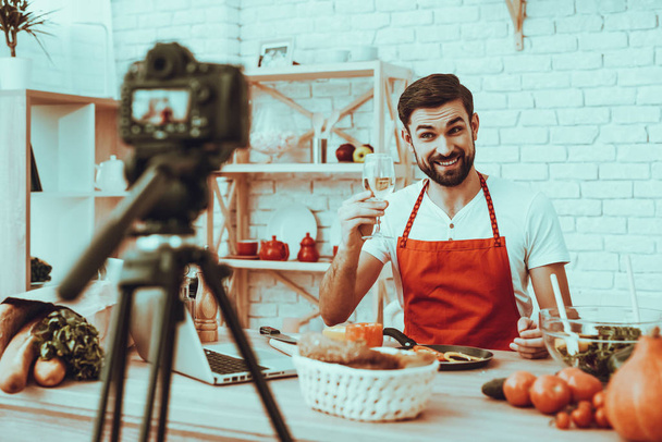 Blogger Makes a Video. Blogger is Smiling Beard Man. Video About a Cooking. Camera Shoots a Video. Laptop and Different Food on Table. Man Showing a White Wine in Wineglass. Man in Studio Interior. - Photo, image
