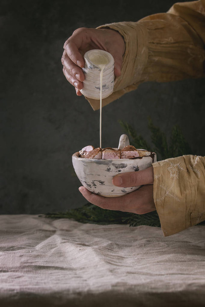 Ceramic cup of hot chocolate with marshmallow s'mores in female hands. Pouring cream from jug. Grey linen table cloth. Winter drink. Dark atmosphere. - Foto, imagen