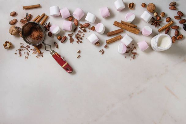Ingredients for hot chocolate. White pink big marshmallow, cocoa beans, cocoa powder, cinnamon, chopped chocolate, nuts over white marble background. Flat lay, space. Sweet food and drink. - Fotografie, Obrázek