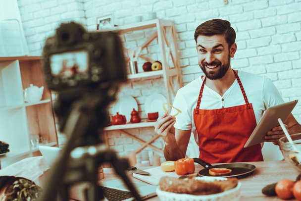 Happy Man Blogger Makes a Video. Video About a Cooking. Camera Shoots a Video. Laptop and Different Food on Table. Man Showing a Pieces of Pepper on Pan. Man Holding a Tablet. Man in Studio Interior. - Foto, Bild