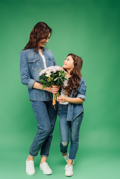 happy mother and daughter in denim looking at each other and holding flowers on green background - Fotografie, Obrázek