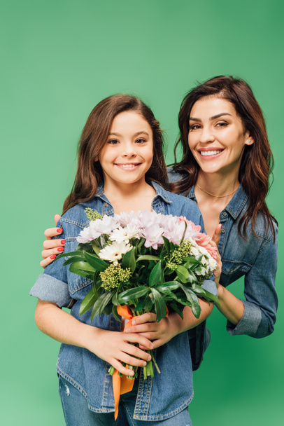 beautiful mother embracing cute daughter with flower bouquet isolated on green - Фото, зображення