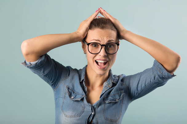 beautiful young girl in a denim shirt and glasses on a blue background with astonishment on her face - Foto, Imagen