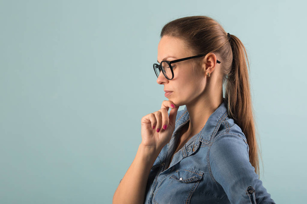 beautiful young girl in a denim shirt and glasses on a blue background with a serious thinking face - Photo, Image