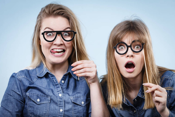 Two happy women holding fake eyeglasses on stick having fun fooling around wearing jeans shirts. Photo and carnival funny accessories concept. - Foto, immagini