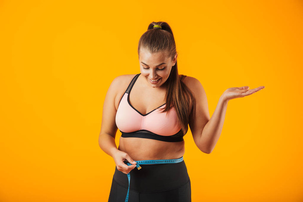 Portrait of a cheerful overweight fitness woman wearing sports clothing standing isolated over yellow background, holding measure tape over her waist - 写真・画像
