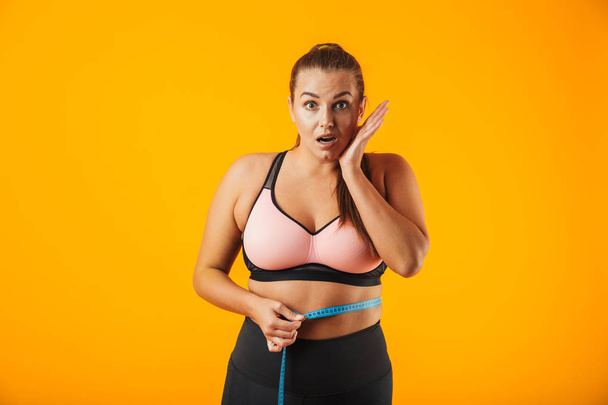 Portrait of excited chubby woman in sportive bra measuring her waist with meter isolated over yellow background - Photo, Image