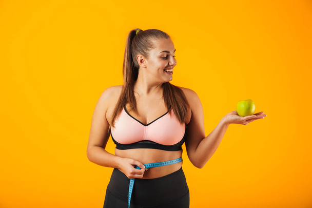 Portrait of caucasian chubby woman in sportive bra measuring her waist with meter and holding apple isolated over yellow background - Foto, Imagen