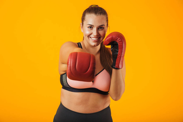 Portrait of joyful big sportswoman in sportive bra wearing boxing gloves practicing isolated over yellow background - Photo, image