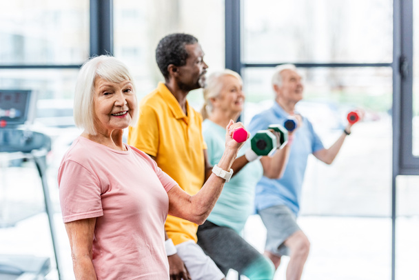 избирательный фокус улыбающейся старшей спортсменки и ее друзей, тренирующихся с гантелями в спортзале
  - Фото, изображение
