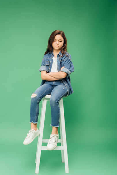 sad child in denim sitting on chair with arms crossed on green background - Photo, Image