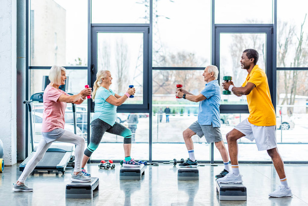 vista lateral de desportistas multiculturais seniores exercício síncrono com halteres em plataformas de degrau no ginásio
 - Foto, Imagem