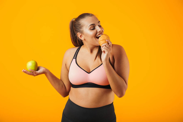 Image of adorable chubby woman in tracksuit holding apple and eating croissant isolated over yellow background - Fotoğraf, Görsel