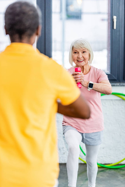 selectieve aandacht van senior vrouw met smartwatch doen oefening met dumbbell op sportschool - Foto, afbeelding
