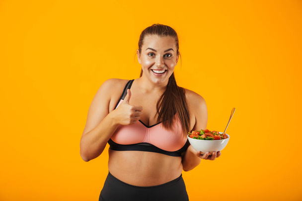 Image of european chubby woman in tracksuit holding plate with green salad isolated over yellow background - Foto, afbeelding