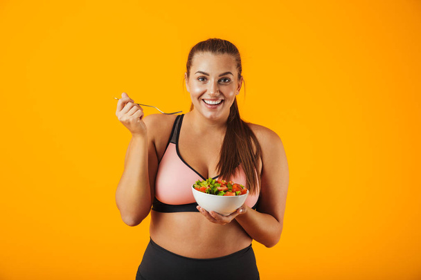 Image of sportive chubby woman in tracksuit eating green salad from plate isolated over yellow background - Foto, afbeelding