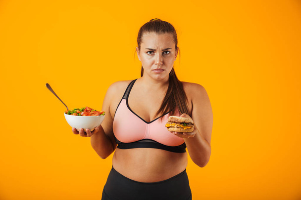 Portrait of an upset overweight fitness woman wearing sports clothing standing isolated over yellow background, holding bowl with salad and a burger - Foto, afbeelding