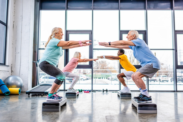 Seitenansicht von glücklichen Senioren multiethnischen Sportlern synchron Kniebeugen auf Trittbrettern in der Turnhalle - Foto, Bild