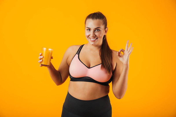 Image of healthy chubby woman in tracksuit smiling and holding glass with orange juice isolated over yellow background - Fotografie, Obrázek
