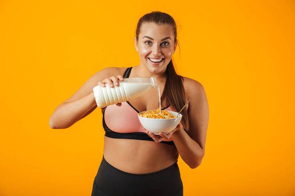 Image of smiling chubby woman in tracksuit holding milk and corn flakes for breakfast isolated over yellow background - Фото, зображення