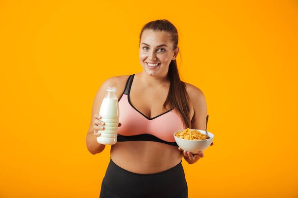Image of european chubby woman in tracksuit holding milk and corn flakes for breakfast isolated over yellow background - Фото, зображення