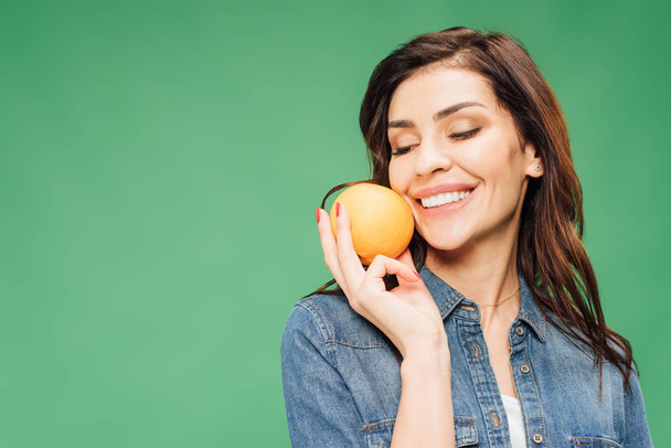 beautiful smiling woman in denim holding orange isolated on green - Foto, afbeelding
