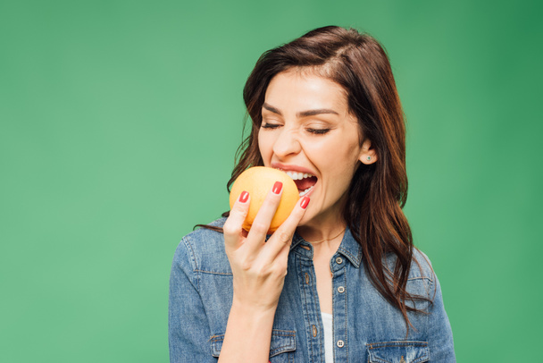 woman in denim biting orange fruit isolated on green - Zdjęcie, obraz