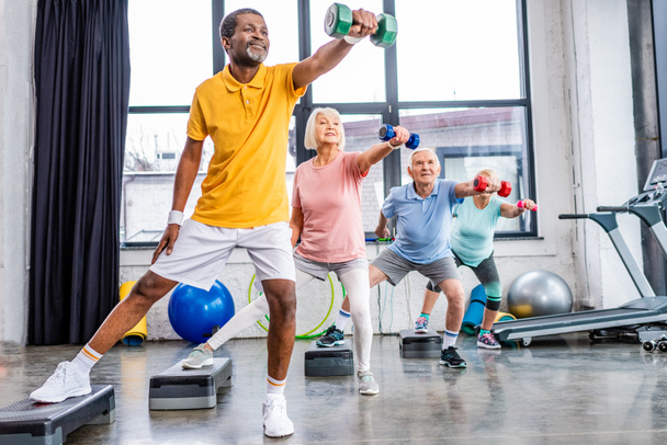 Senioren trainieren synchron mit Hanteln auf Trittbrettern im Fitnessstudio - Foto, Bild