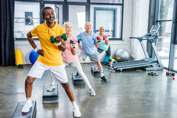 deportistas senior multiculturales ejercicio sincrónico con mancuernas en plataformas escalonadas en el gimnasio
 - Foto, imagen