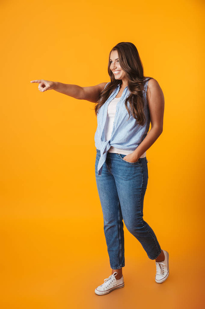 Portrait of a cheerful young casual woman standing isolated over yellow background, pointing fingers away at copy space - Photo, Image